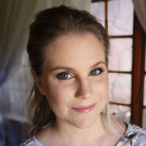 Close-up of a woman indoors with blue eyes, light brown hair pulled back, near window with wooden frames.