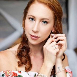 Woman with red curly hair and blue eyes adjusting an earring wearing floral off-shoulder top.