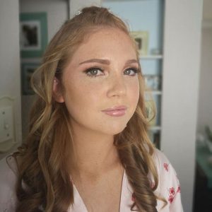 Woman with wavy light brown hair, makeup, and floral-patterned light-colored top indoors.