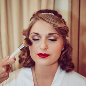 Woman with styled hair and red lipstick having makeup applied on her cheek with a brush.