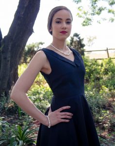 Woman in sleeveless black dress with pearl necklace and bracelet posing outdoors in garden.
