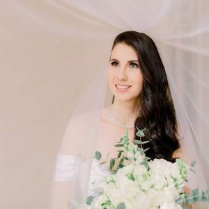 Bride with long dark hair in white off-shoulder dress and veil holding a bouquet of white flowers.
