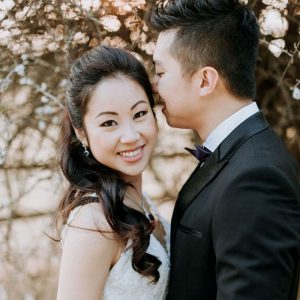 Couple dressed in wedding attire posing outdoors with blooming branches in the background.
