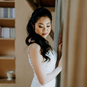 Woman in white lace dress standing by a curtain with eyes closed and blurred bookshelf background.