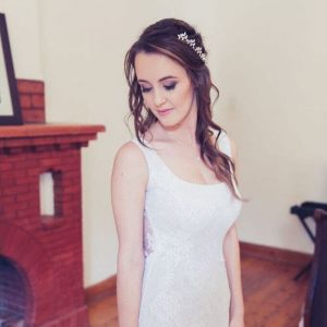 Woman in white lace sleeveless dress standing indoors near a brick fireplace and wooden floor.
