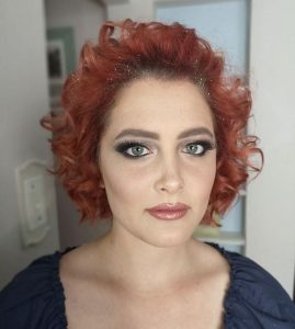 Close-up portrait of a person with curly red hair, glitter, dramatic eye makeup, and glossy lips.