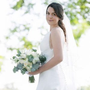 Bride in white wedding dress and veil holding a white flower and greenery bouquet outdoors.