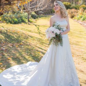 Bride in strapless white wedding gown holding bouquet outdoors on a sunny day with autumn leaves.