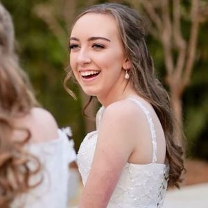 Young woman in a white dress with pearl earrings smiling outdoors with blurred greenery background.