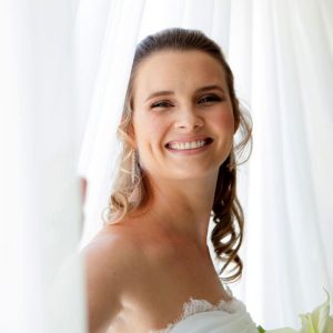 Smiling woman in a strapless white dress holding white flowers standing by sheer curtains